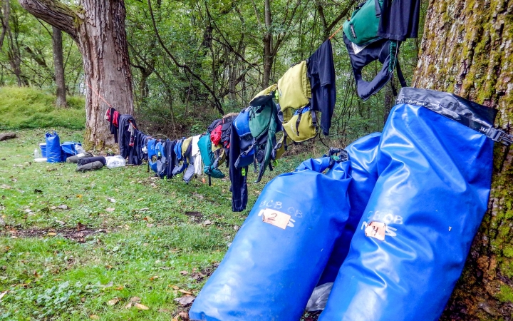 A clothesline tied between two trees holds gear that is drying. There are two dry bags resting against the tree in the foreground. 